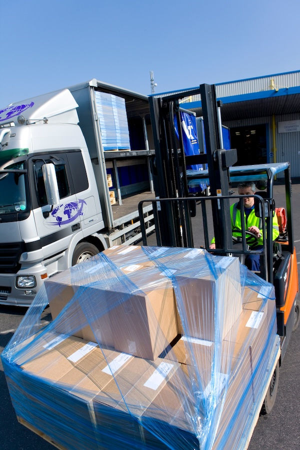 A forklift operator moves a pallet of boxes wrapped in blue plastic outside a loading dock. A truck with open cargo holds is nearby.