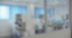 Scientists in lab coats work in a bright laboratory. One uses a microscope, another handles equipment. Blue blinds, sterile environment.