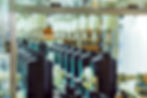 Conveyor with black bottles being filled by a machine in a factory. Bright lighting, metal and plastic components are visible. Industrial setting.