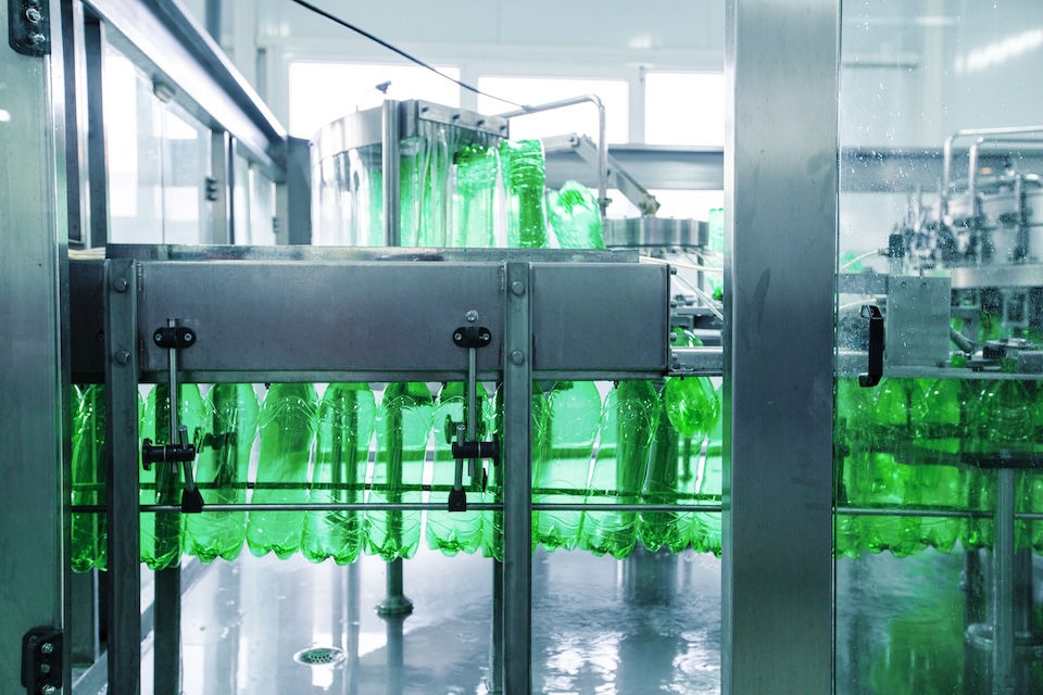 Green plastic bottles moving through a stainless steel conveyor system in a factory. Bright, clean industrial setting with glass windows.