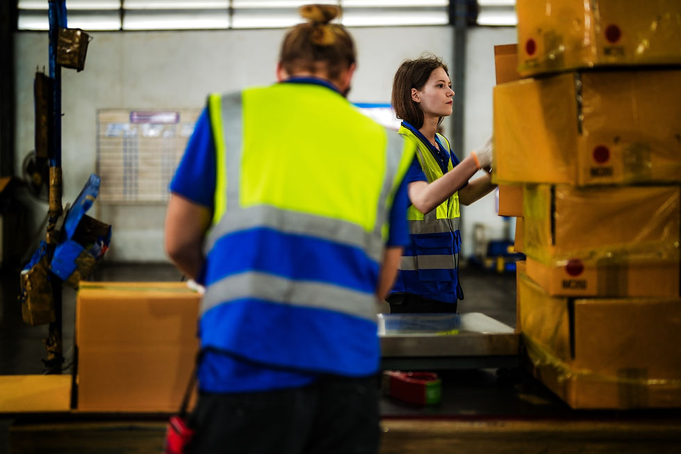 Two warehouse workers in yellow vests handle stacked boxes in an industrial setting. The background includes a board with blurred text.