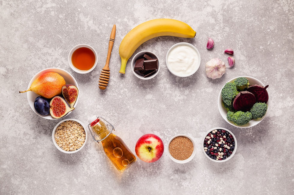 Assorted foods on a gray surface, including a banana, yogurt, vegetables, fruits, honey, oats, and a bottle of amber liquid, arranged artistically.