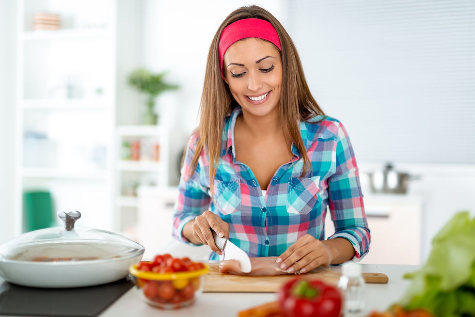 Woman in a plaid shirt and red headband chops chicken on a cutting board, smiling in a bright kitchen with fresh vegetables nearby.