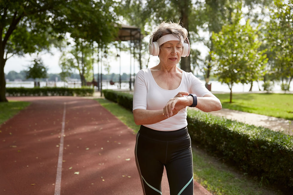 Elderly woman on a track checks smartwatch, wearing headphones and a headband. Sunny park setting, visible trees, and lush greenery.
