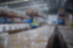 Warehouse with two workers in safety vests and helmets inspecting shelves of boxes. Blue metal racks, high ceiling, bright lighting.