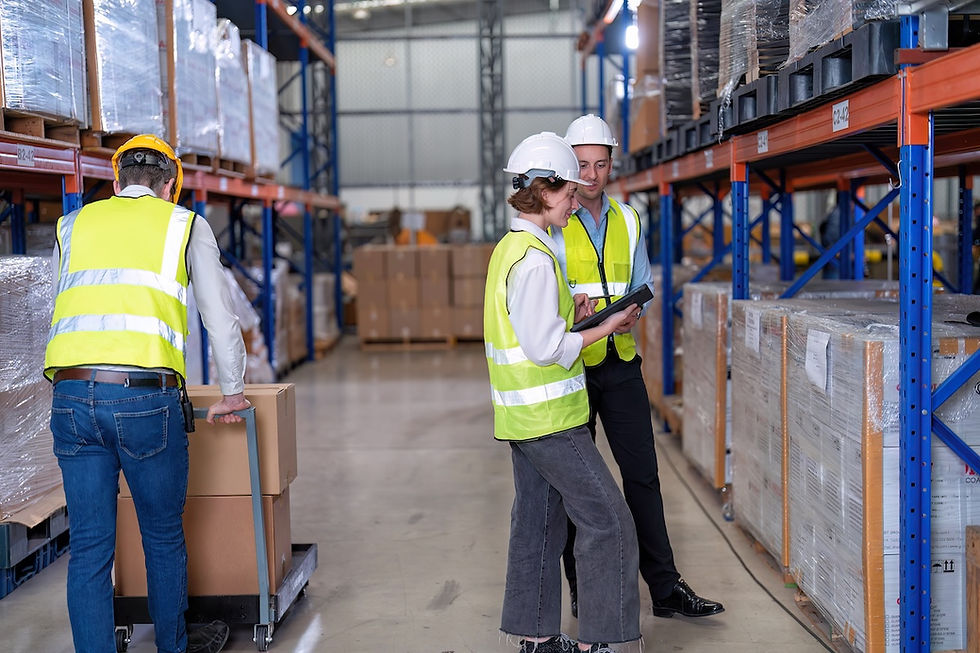 Warehouse scene with two workers in vests and helmets discussing a tablet. Another person pushes a cart with boxes. Shelves of packages line the aisle.