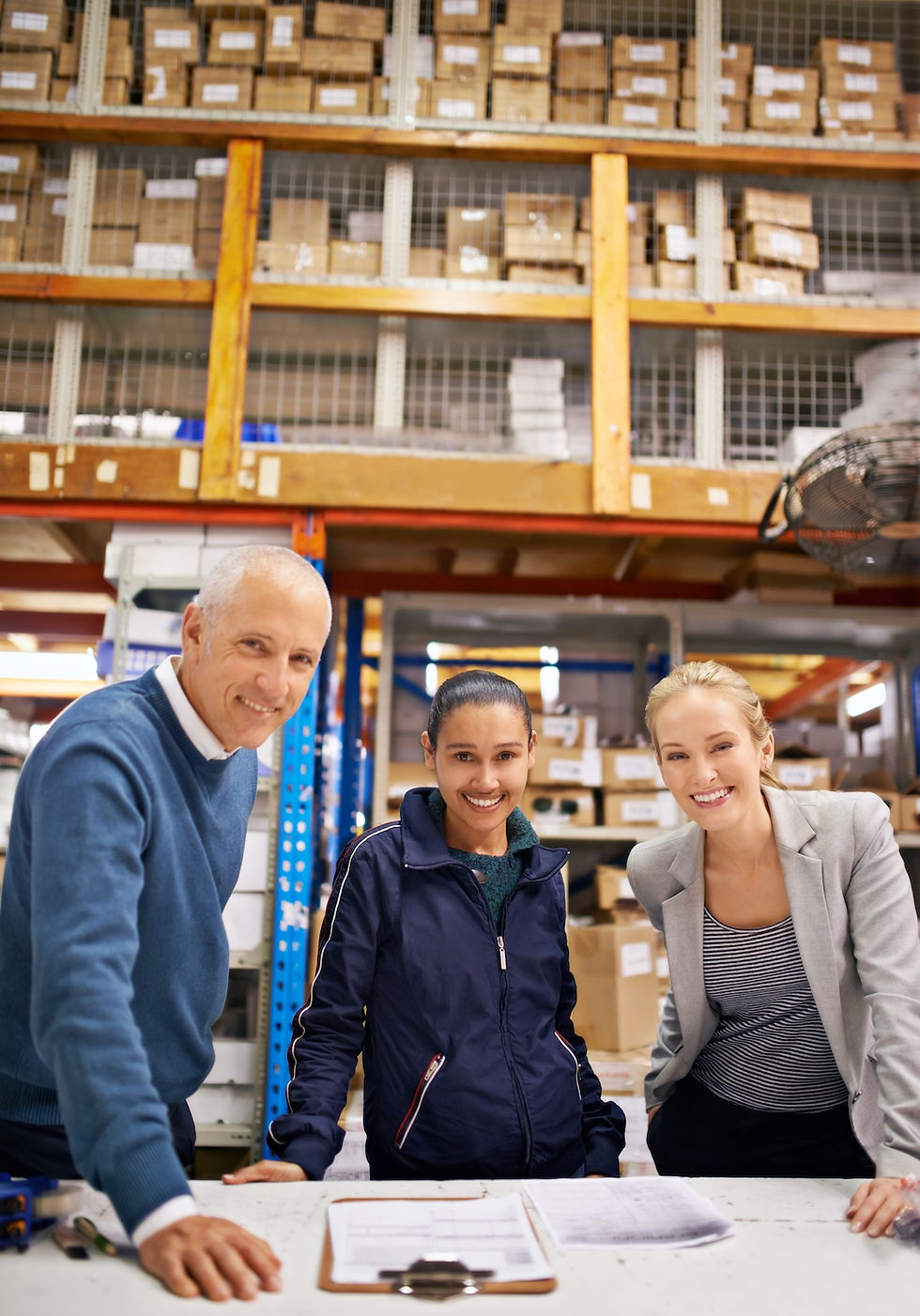 Three smiling people in a warehouse with shelves of boxes. They stand around a table with papers, suggesting teamwork and productivity.