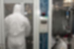Person in white hazmat suit stands in a sterile lab. There's a device labeled "Negative Pressure Indicator" with buttons and cables.