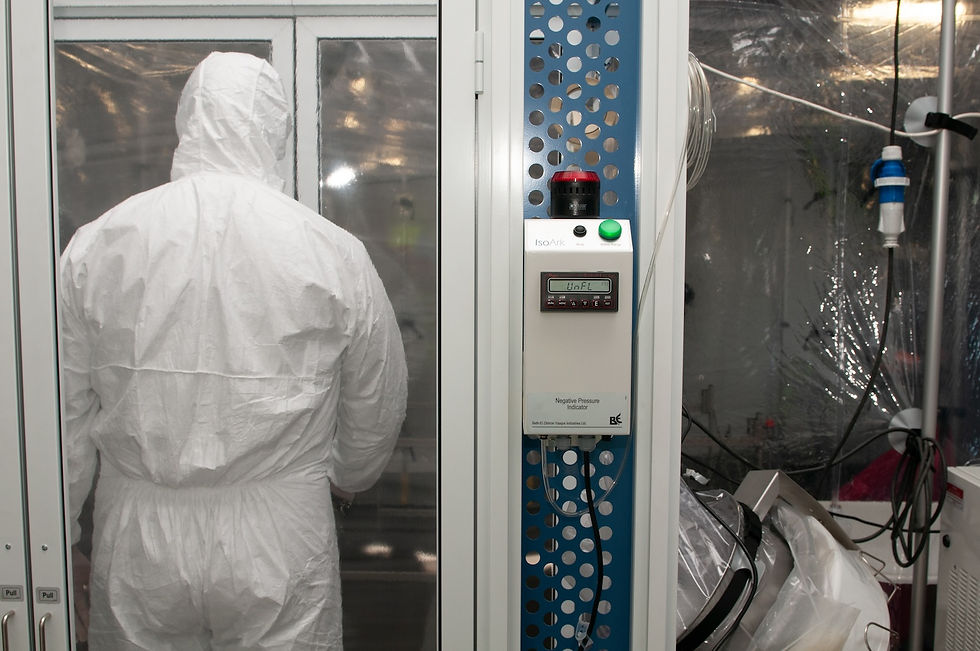 Person in white hazmat suit stands in a sterile lab. There's a device labeled "Negative Pressure Indicator" with buttons and cables.