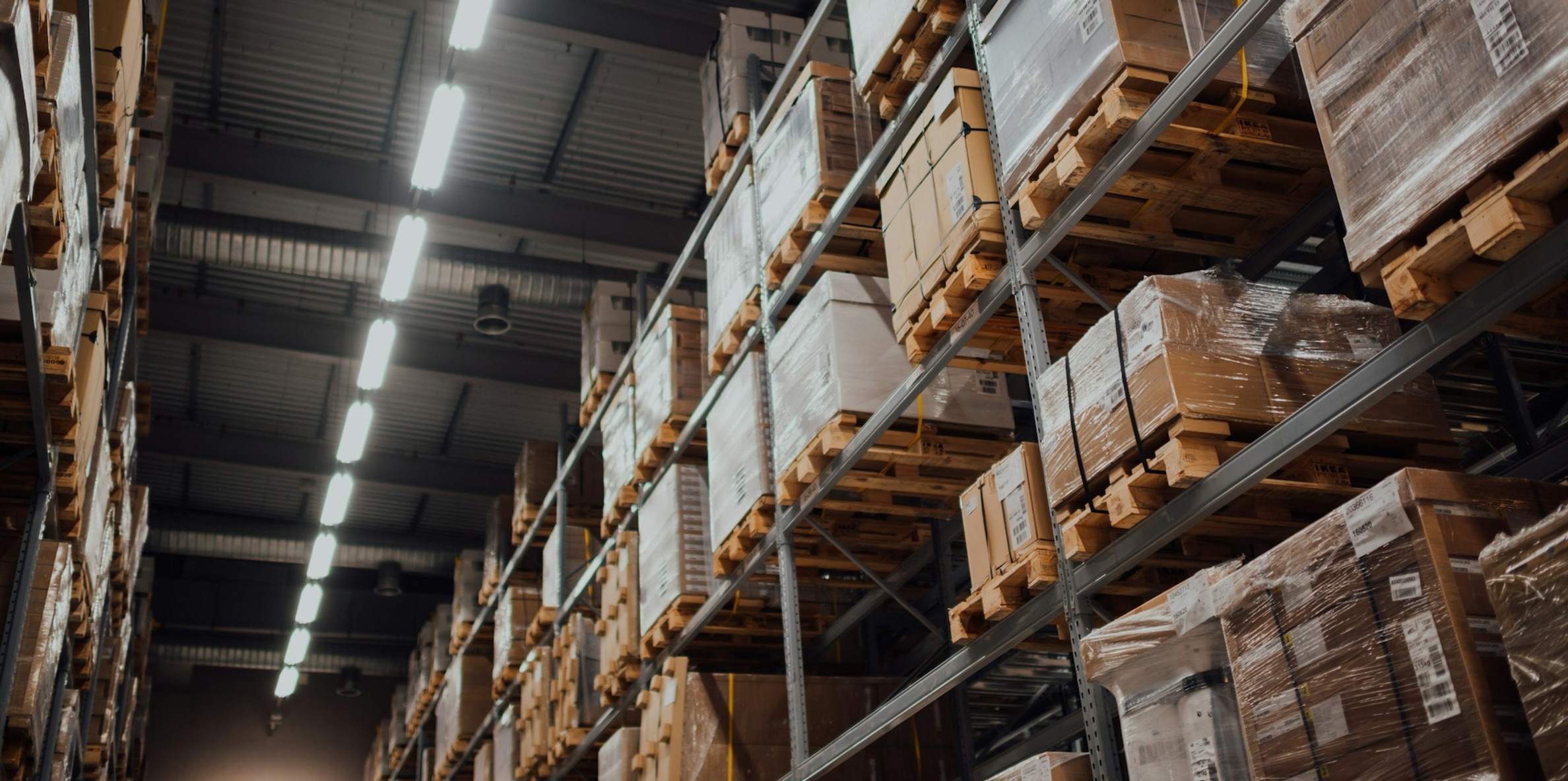 Tall warehouse racks filled with shrink-wrapped pallets and boxes under bright fluorescent lights, no people visible