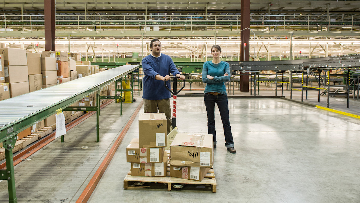 Two warehouse workers stand by a pallet jack loaded with labeled boxes in a large distribution center with conveyor belts.