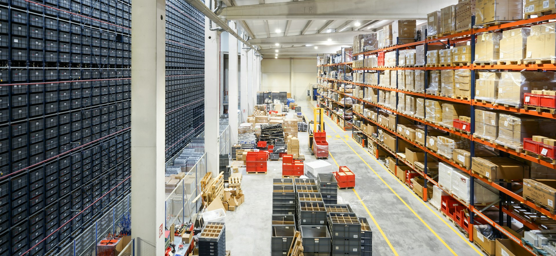 Bright warehouse aisle with tall orange racks of boxed goods and rows of black crates under concrete beams.