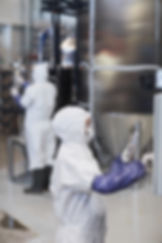 People in white hazmat suits work in a clean industrial setting. One uses a touchscreen device. Shiny metal tanks in the background.