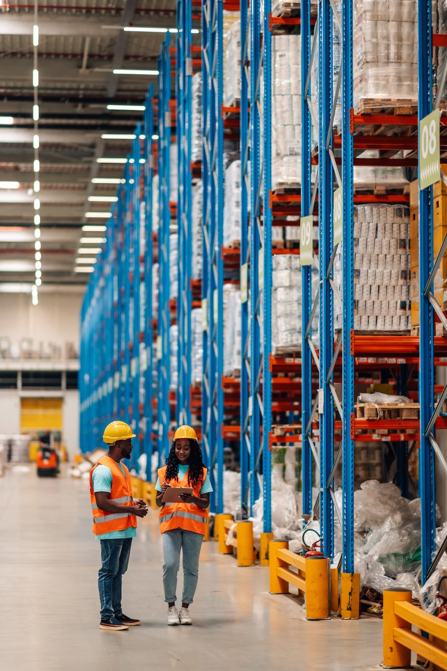 Two workers in orange vests and yellow helmets converse in a warehouse aisle lined with blue and orange shelves. One holds a clipboard.