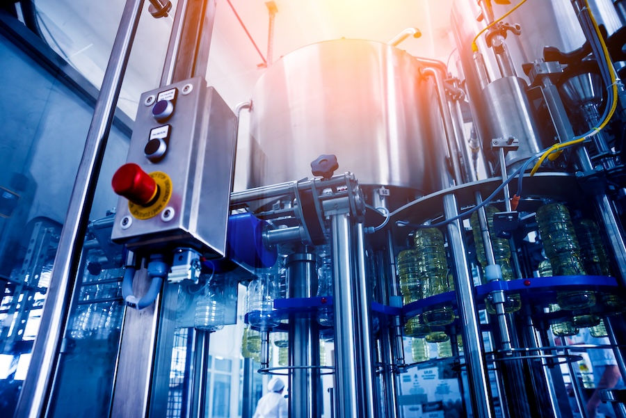 Industrial bottling machine filling plastic bottles in a factory. Stainless steel tanks and control panel visible. Bright, metallic setting.