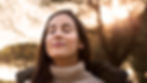 Woman in a beige turtleneck with a serene expression stands outdoors at sunset, eyes closed, with soft sunlight filtering through trees.