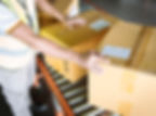 Worker in a vest moves large brown boxes on a conveyor belt in a warehouse. Labels are visible on the boxes, indicating shipping.