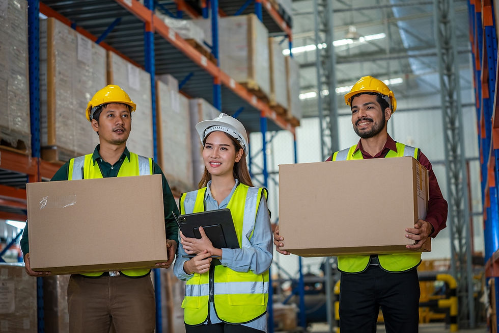 Workers in safety vests and helmets stand in a warehouse. Two hold boxes; one holds a tablet, smiling. Shelves filled with boxes in the background.