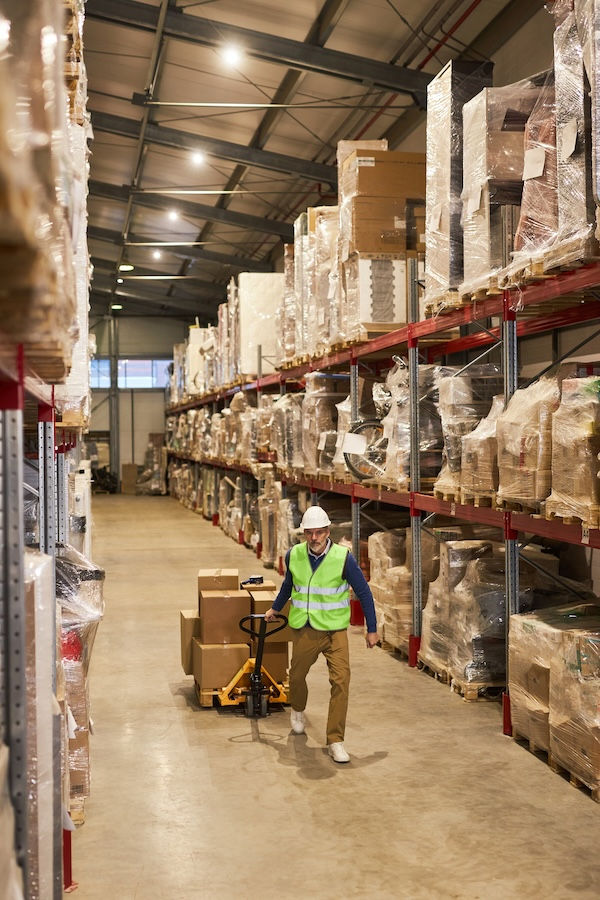 Warehouse worker in a high-vis vest and helmet moves boxes with a pallet jack. Stacked shelves surround in a spacious, well-lit area.