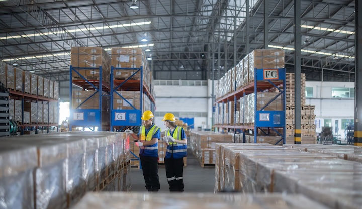 Warehouse with two workers in yellow helmets and vests checking boxes. Large shelves filled with pallets. Bright lighting, industrial setting.