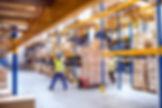 Workers in safety vests and helmets move boxes with a pallet jack in a large warehouse with yellow shelving and stacked goods.