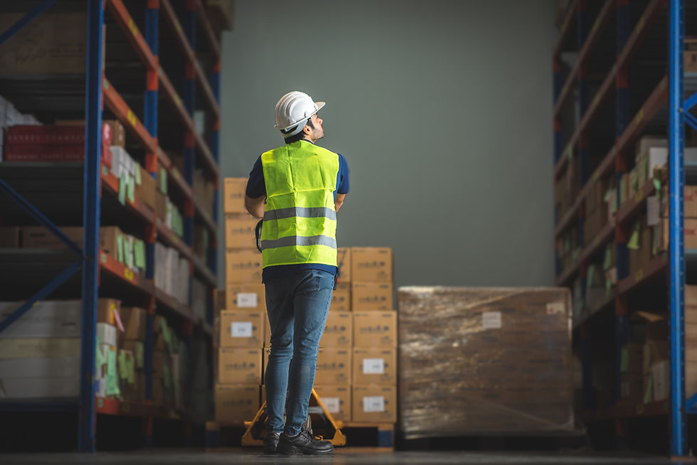 Worker in yellow vest and hard hat observes stacked boxes on shelves in a warehouse. He stands beside a pallet jack, appearing focused.