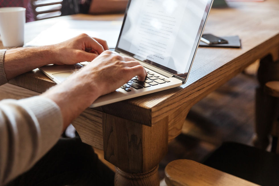 Hands typing on a laptop at a wooden table, with a cup and phone nearby. The screen shows text, suggesting a focused work environment.