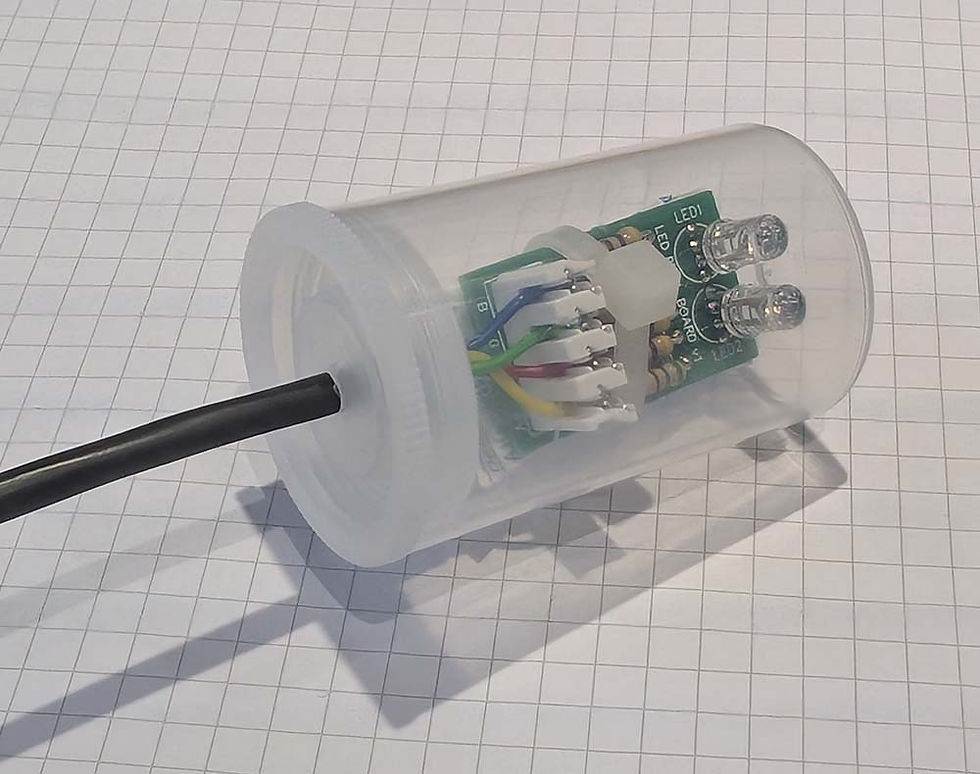 Transparent plastic cylinder with electronic circuit and wires inside, resting on graph paper. Contains LEDs, green circuit board, and black cable.