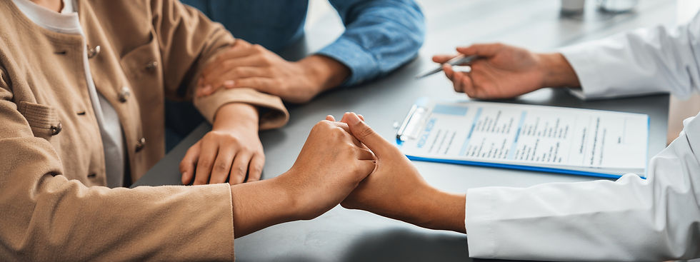 A supportive doctor holds the hands of a couple during a consoling conversation, with a medical chart visible on the desk.