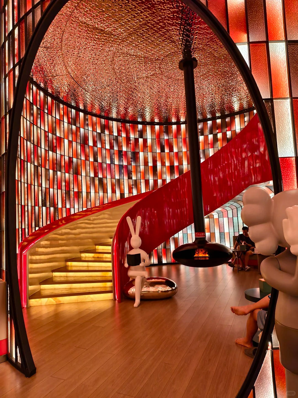 Colorful room with red and orange checkered walls, a glowing red spiral staircase, and a white bunny statue holding a book. Cozy seating area.