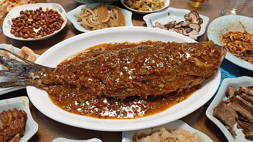A glazed fish in spicy brown sauce on a white platter, surrounded by various side dishes like peanuts, mushrooms, and sliced meats.