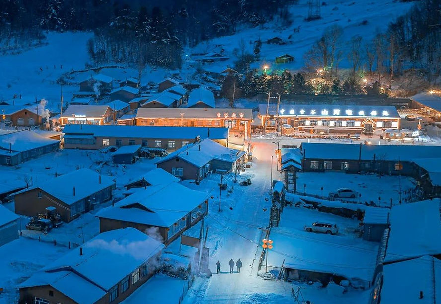 Snowy village at night with warm lights, surrounded by trees. Three people walk along a snowy road. Buildings display lit signs, adding charm.
