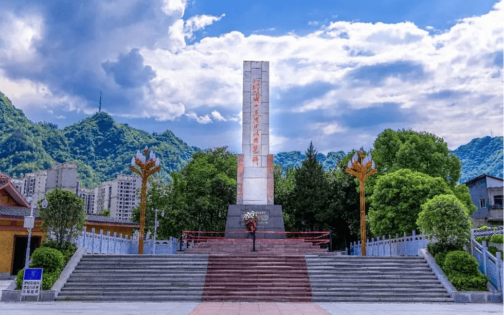 Monument with red inscriptions on a platform, set against green hills and a cloudy sky. Buildings and trees surround the area.