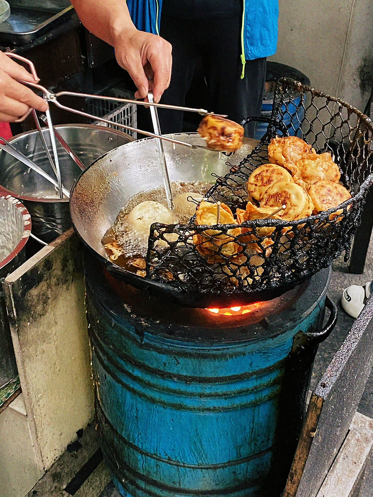 Hands use tongs to fry pastries in a silver pan over a blue barrel. Golden pastries rest on a mesh rack. Street food scene.