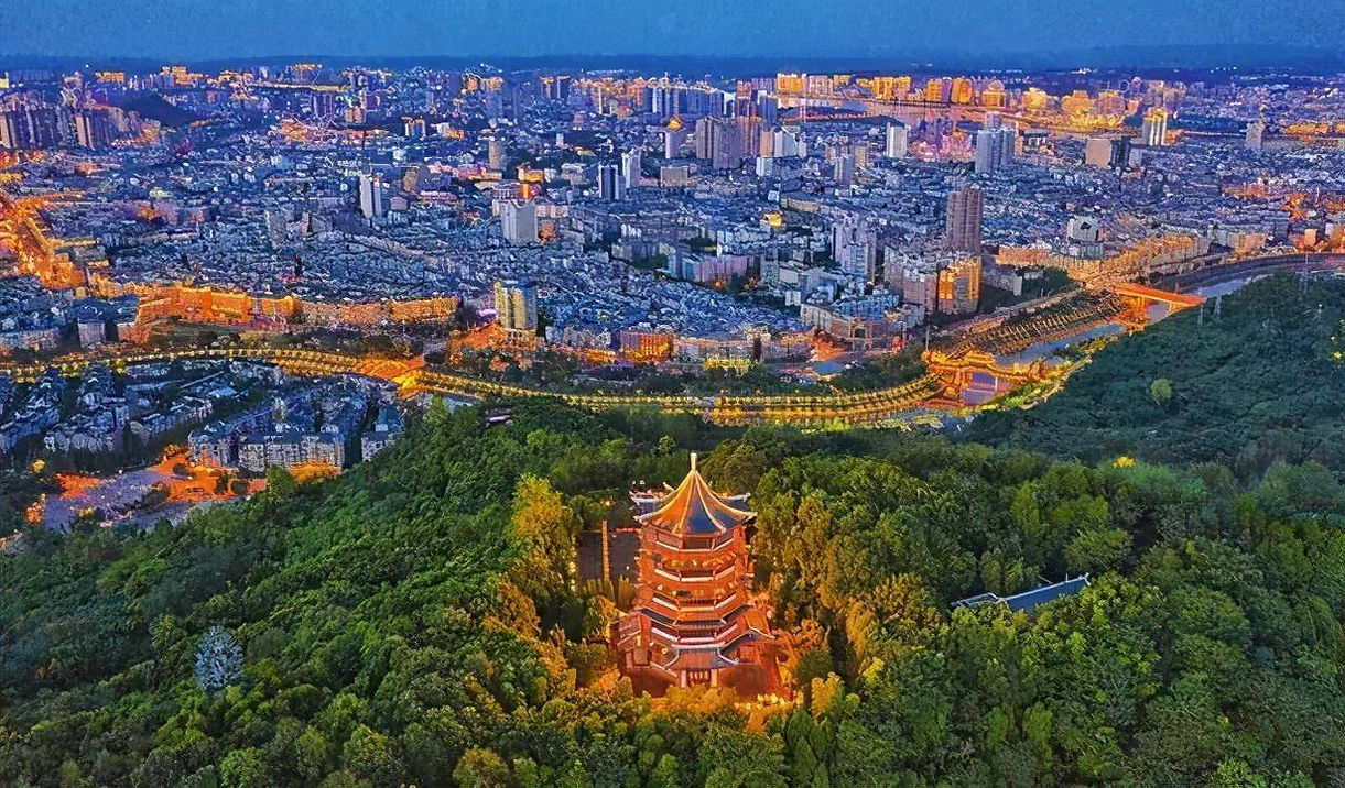 Aerial view of a brightly lit city at dusk, with a pagoda in a lush green forest in the foreground. Buildings glow with warm lights.