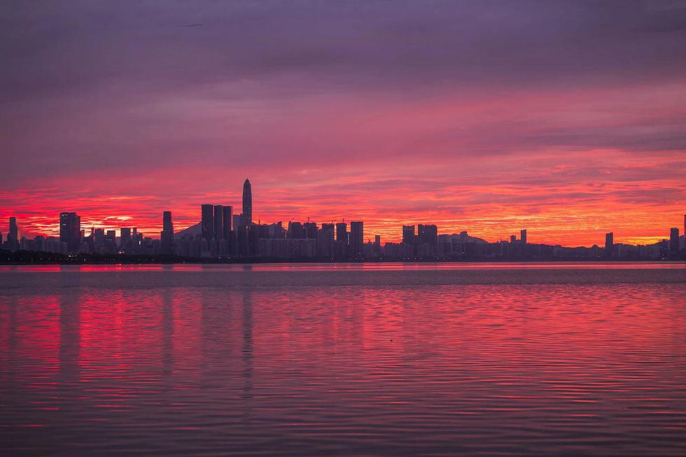 City skyline at sunset with vibrant red and purple hues reflecting on calm water. Buildings silhouetted against dramatic sky.
