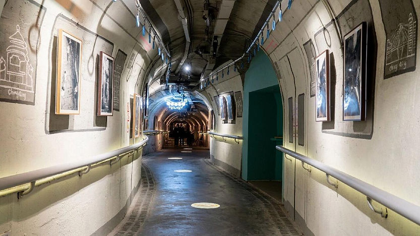 Underground tunnel with artworks on walls, lit by neon ceiling lights. Dark floor with circular patterns; people visible in the distance.