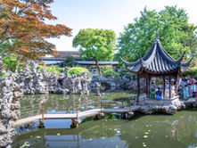 People gather at a traditional Chinese pavilion by a pond, surrounded by lush greenery and rock formations. The scene is tranquil and vibrant.
