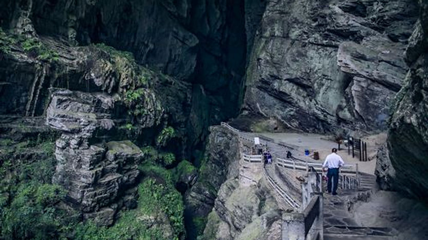 People walking along a path inside a large, rocky cave with green moss. The setting is remote and peaceful, and there's no visible text.