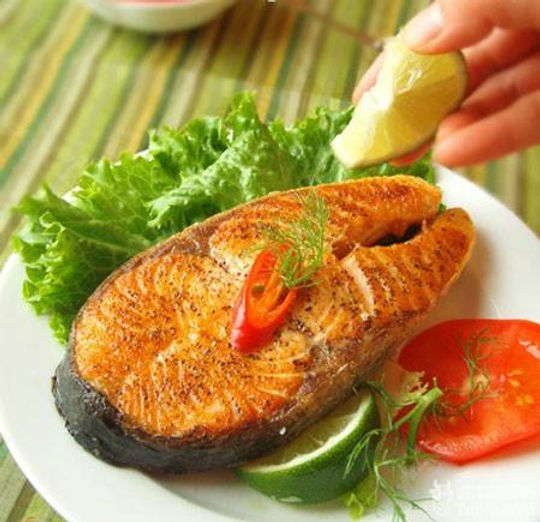 Grilled salmon steak on a plate with lettuce, lime, and tomato. A hand squeezes lime above. Green striped tablecloth background.