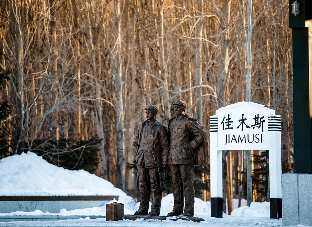 Bronze statues of two men in uniforms stand beside a sign reading "JIAMUSI" with snow and bare trees in the background.