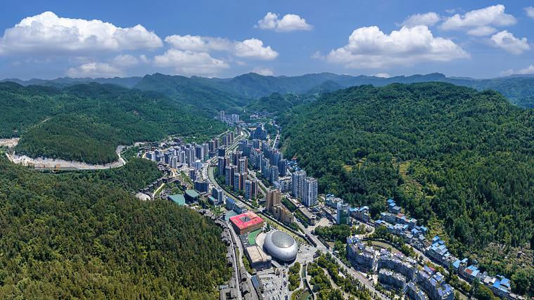 Aerial view of a city nestled in lush green mountains, featuring rows of buildings and a distinctive dome structure under a partly cloudy sky.