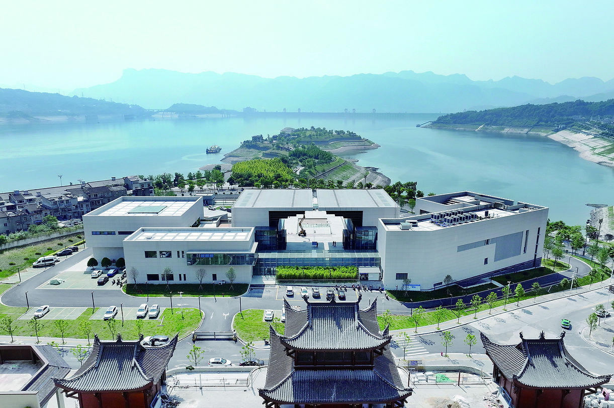 Modern white building near a river with mountain backdrop. Traditional rooftops in foreground. Bright and clear sky sets a peaceful mood.