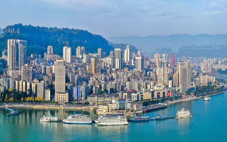Cityscape with tall buildings along a riverbank. Several white boats float on the calm blue water. Hills in the background under a clear sky.