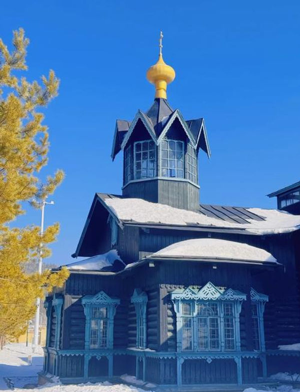 Blue wooden church with a golden onion dome and intricate trim, set against a bright blue sky. Snow covers roof and ground. Peaceful scene.