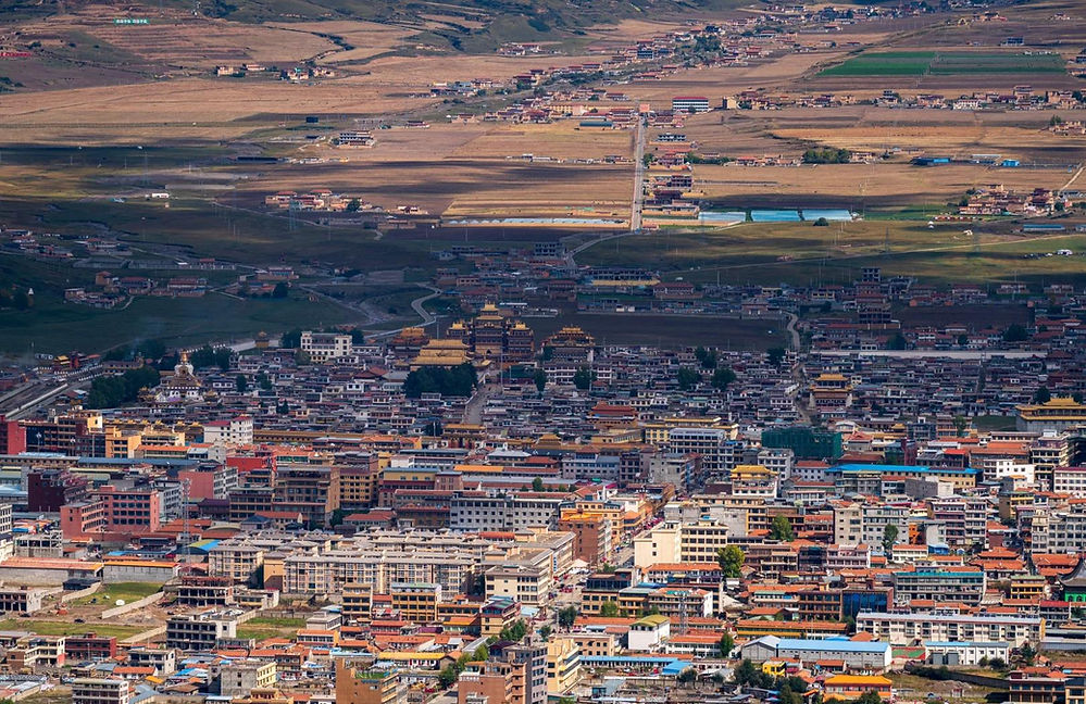 Aerial view of a densely built town with colorful buildings, a temple, and fields in the background under a partly cloudy sky.