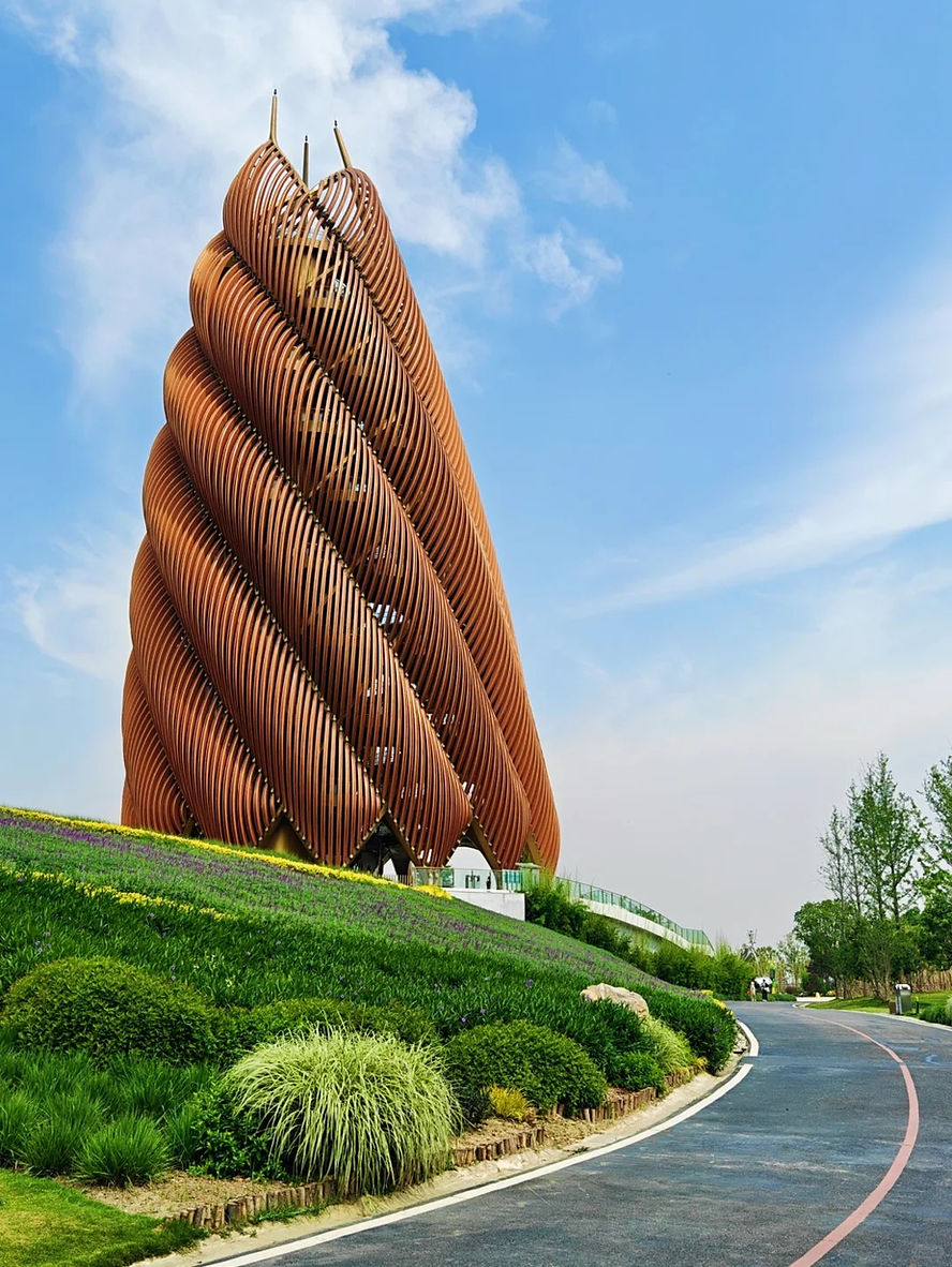 Futuristic brown spiral building in a landscaped park with lush greenery and a winding path, set against a clear blue sky.