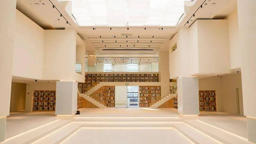 Bright, spacious library with high ceiling and skylights. White interior, symmetrical stairs, and bookshelf-lined walls create a calm atmosphere.