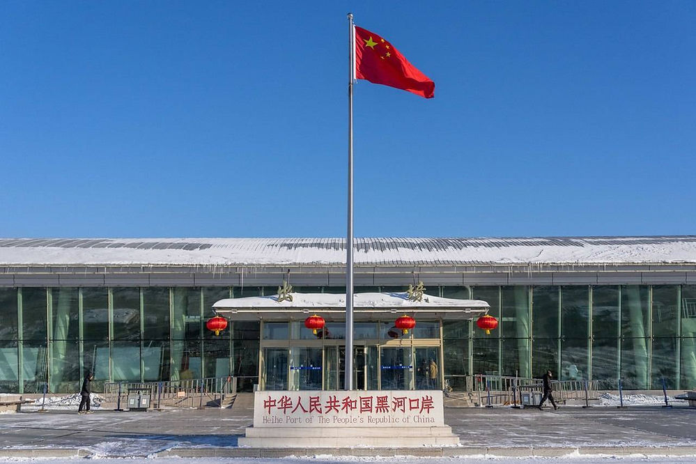 Red Chinese flag flies outside Heihe Port building in snow. Glass facade, red lanterns, and sign in Chinese and English under clear blue sky.