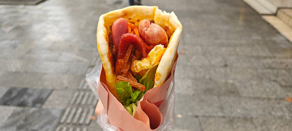 Close-up of a stuffed pita wrap with lettuce, fried egg, sausages, and vegetables, held against a gray pavement background.